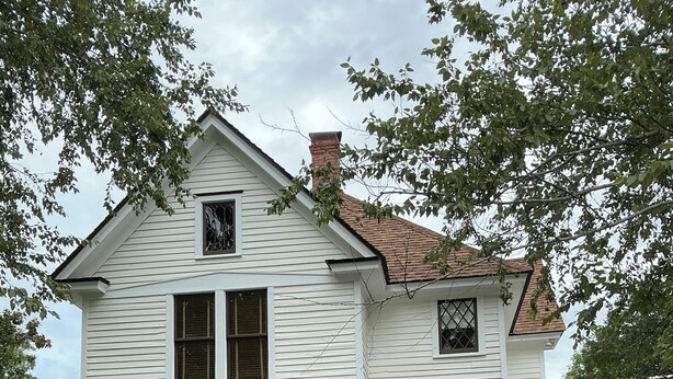 Een wit huis met twee verdiepingen, een bakstenen schoorsteen, een veranda met een hangstoel en een tegelpad dat naar de ingang leidt.