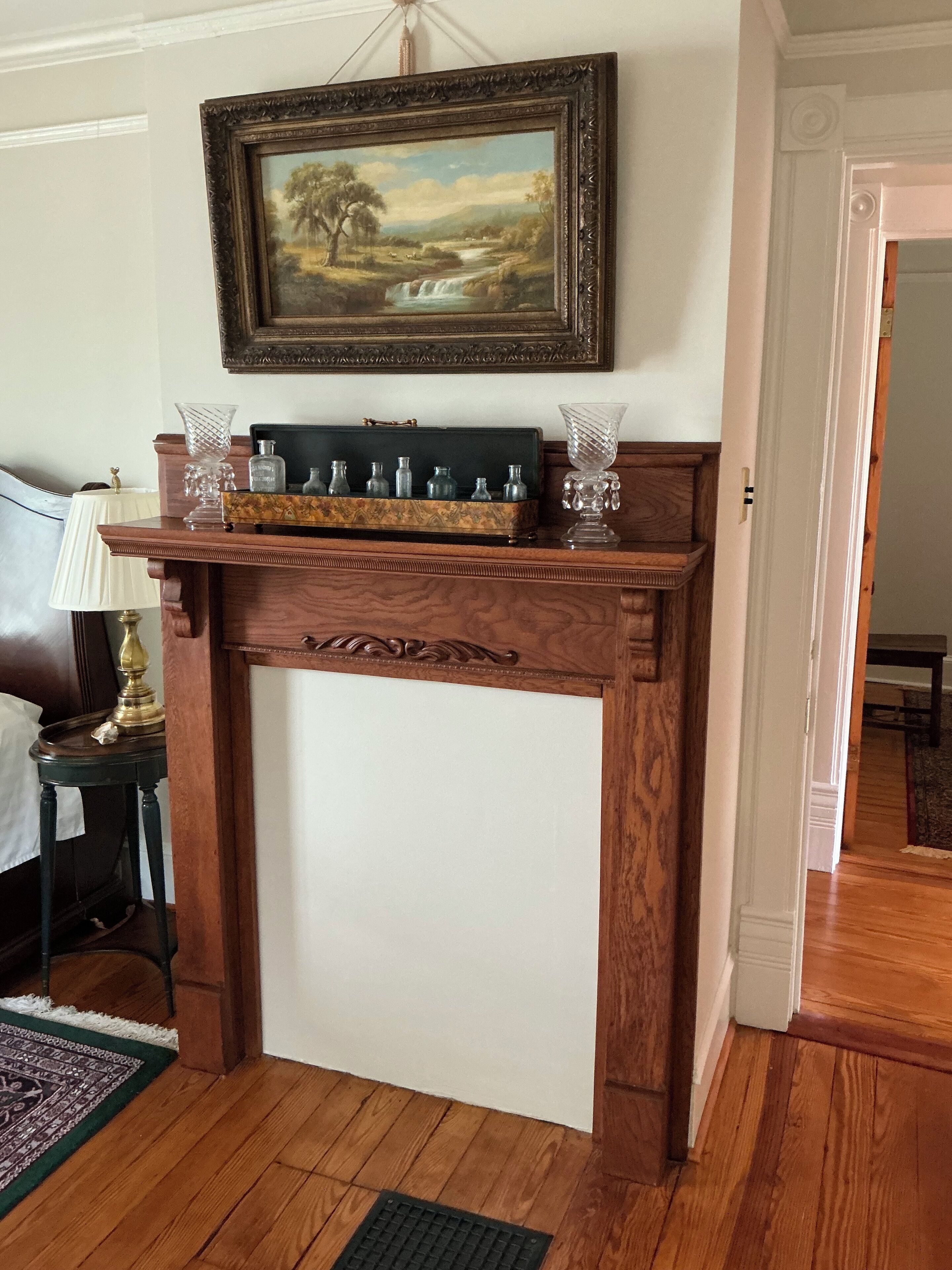 Uma lareira de madeira com mantelo que exibe uma pintura paisagística emoldurada, um vaso e uma caixa decorativa.