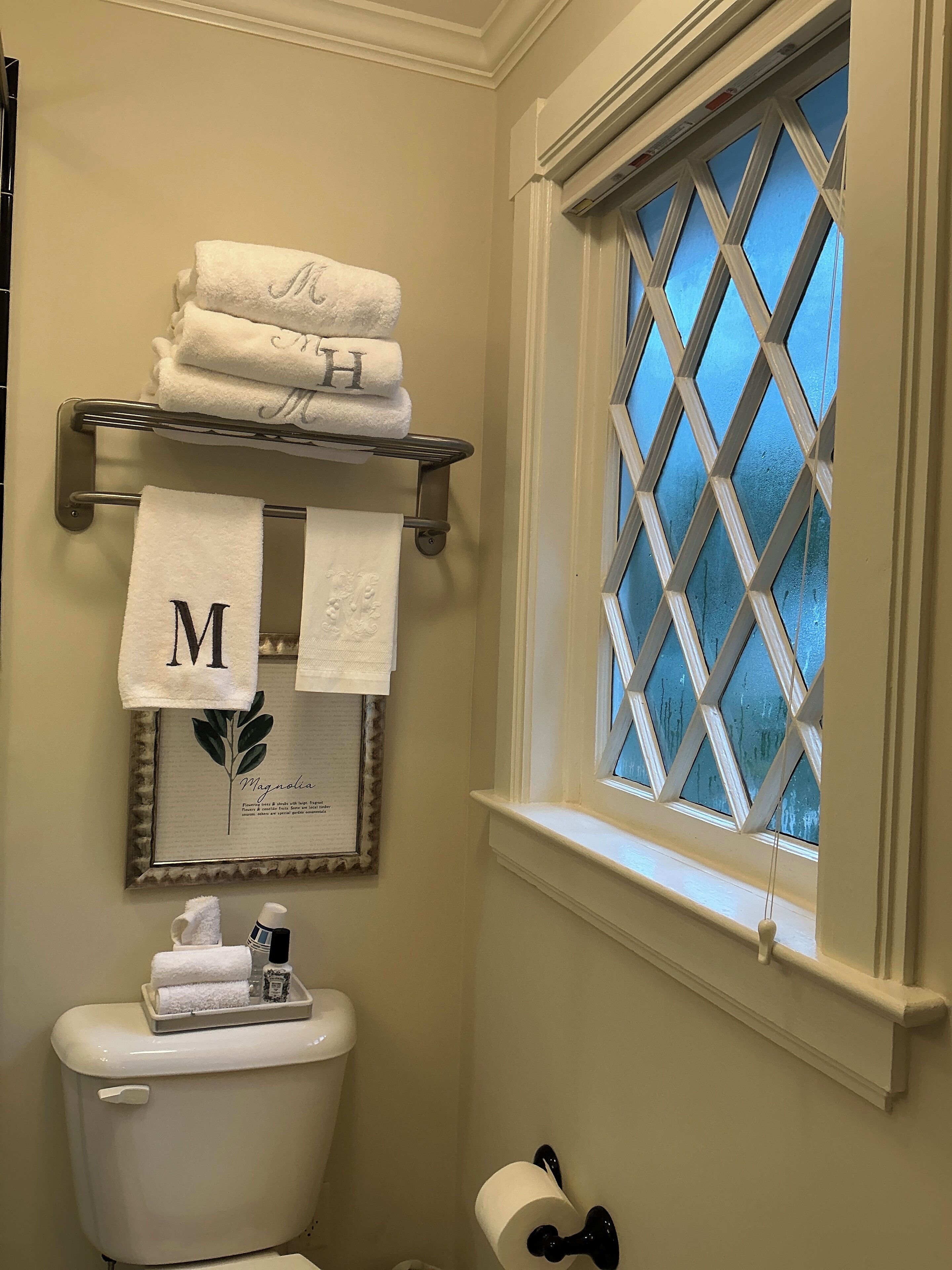 A bathroom with a toilet, a window with a diamond pattern, a towel rack with towels, and a framed picture on the wall.