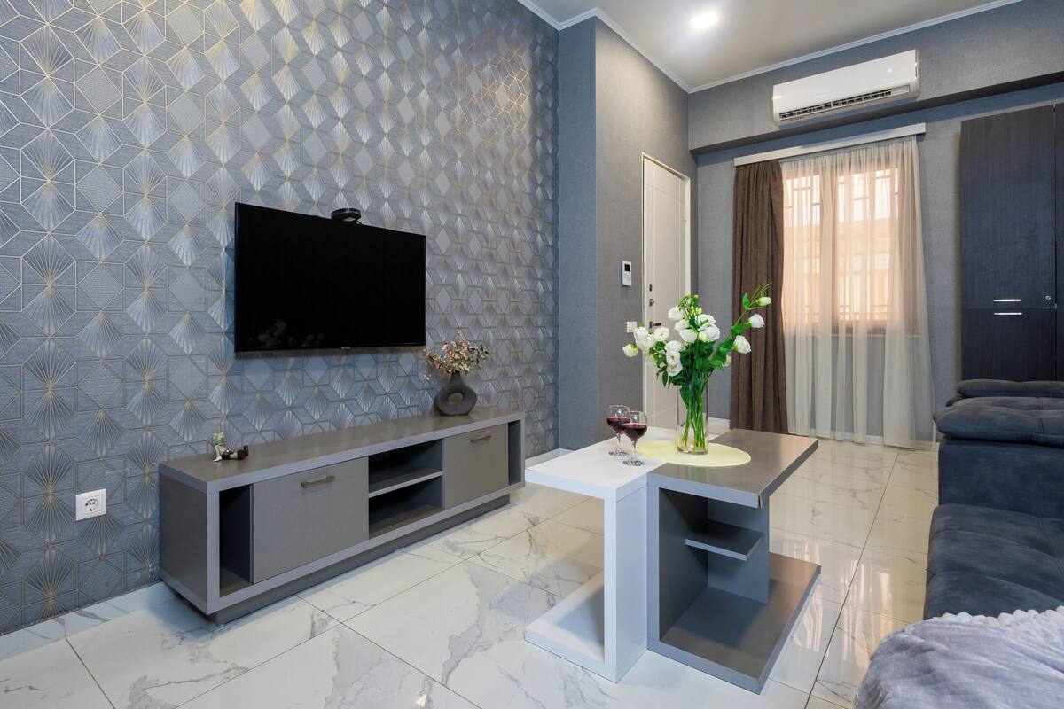 Una sala moderna con un televisor de pantalla plana montado en una pared texturizada, un mueble de entretenimiento gris, una mesa de centro blanca con un jarrón de flores y un sofá gris.