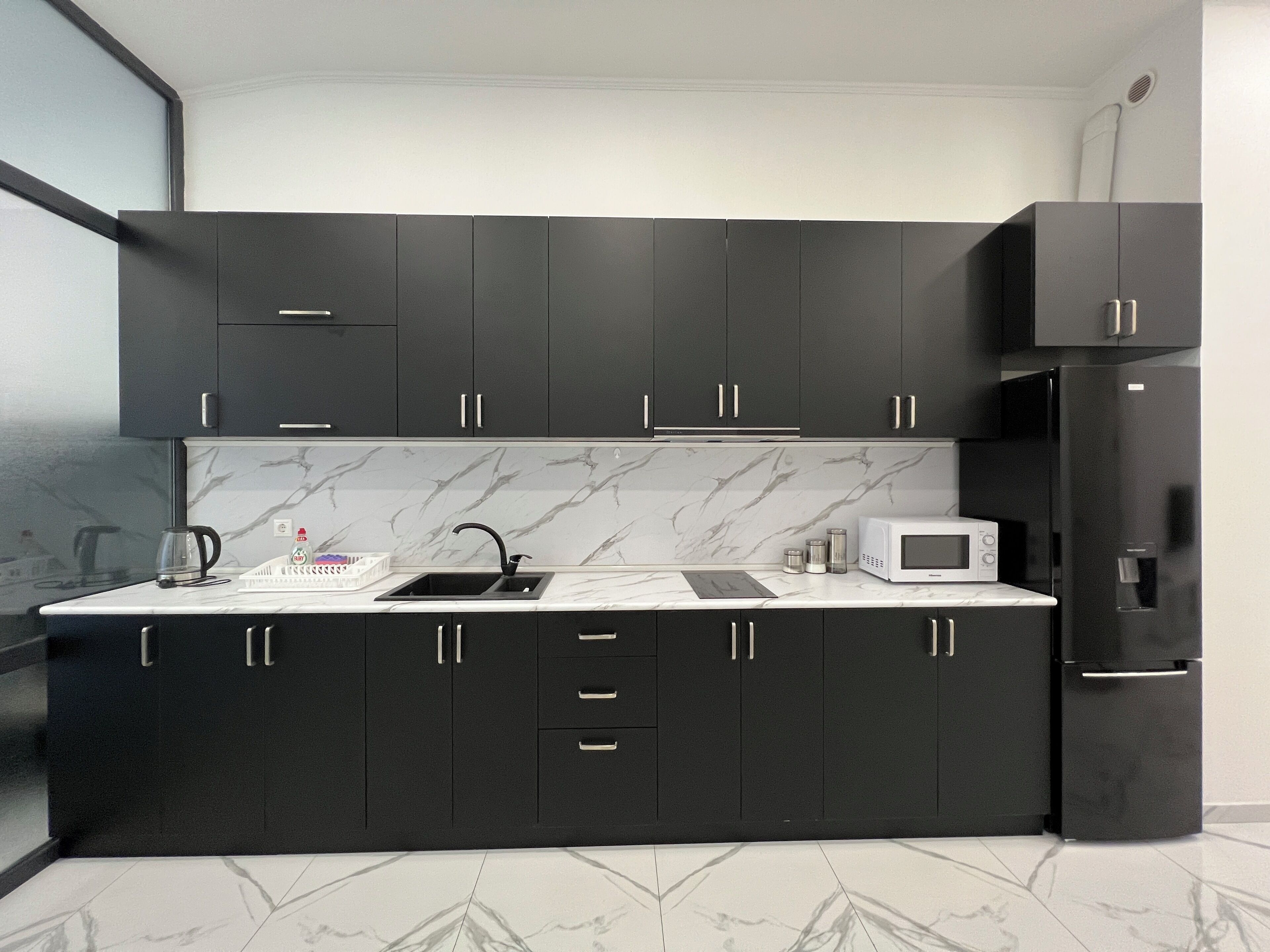 Une cuisine moderne avec des meubles de couleur sombre, un plan de travail blanc, un évier, une bouilloire, un four à micro-ondes et un réfrigérateur.