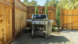 A portable Weber grill with a side burner and a cart, set up in a backyard with wooden fencing and palm trees. - Beautiful Turn of the Century Home with Pet Friendly Yard (Martinez)