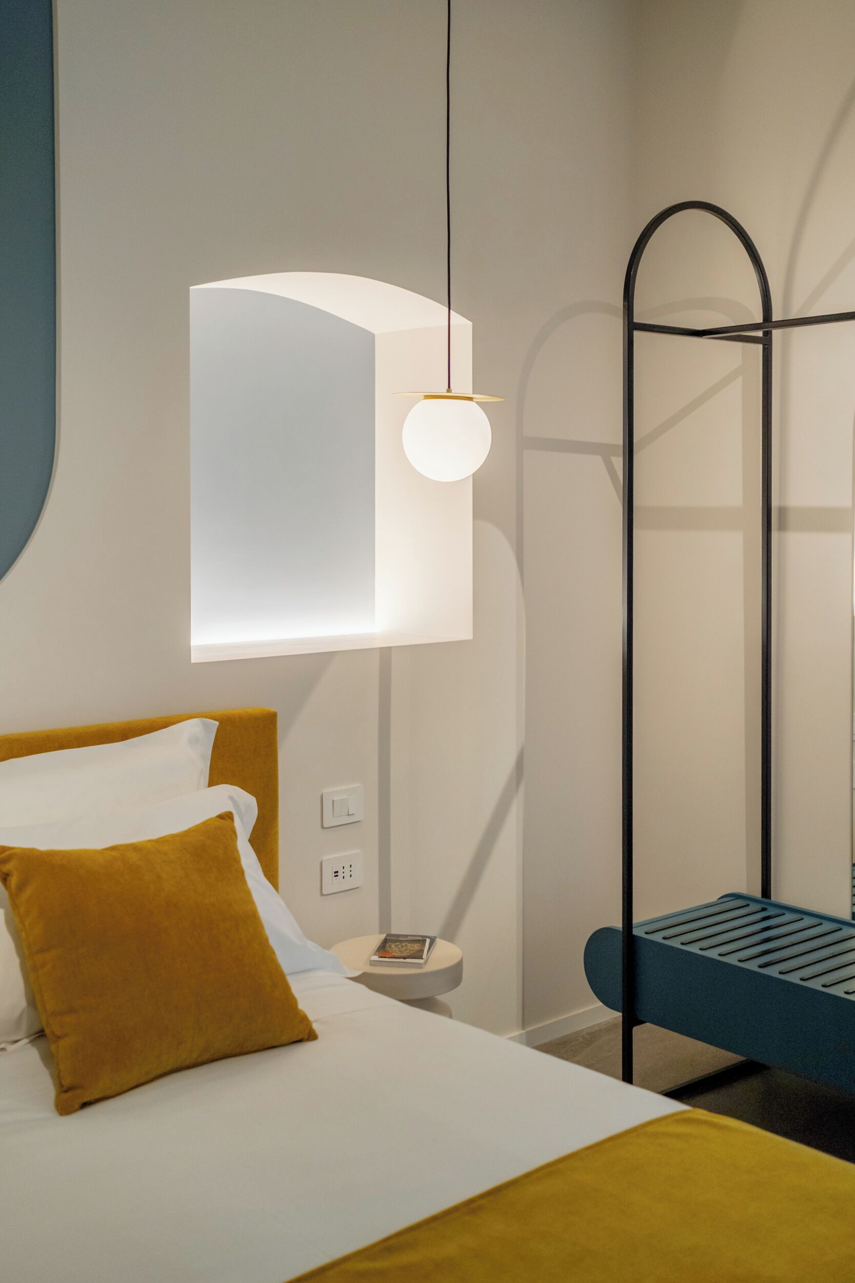 Une chambre d’hôtel moderne comprenant un lit, une table de chevet, une lampe de lecture et une structure originale en métal noir.