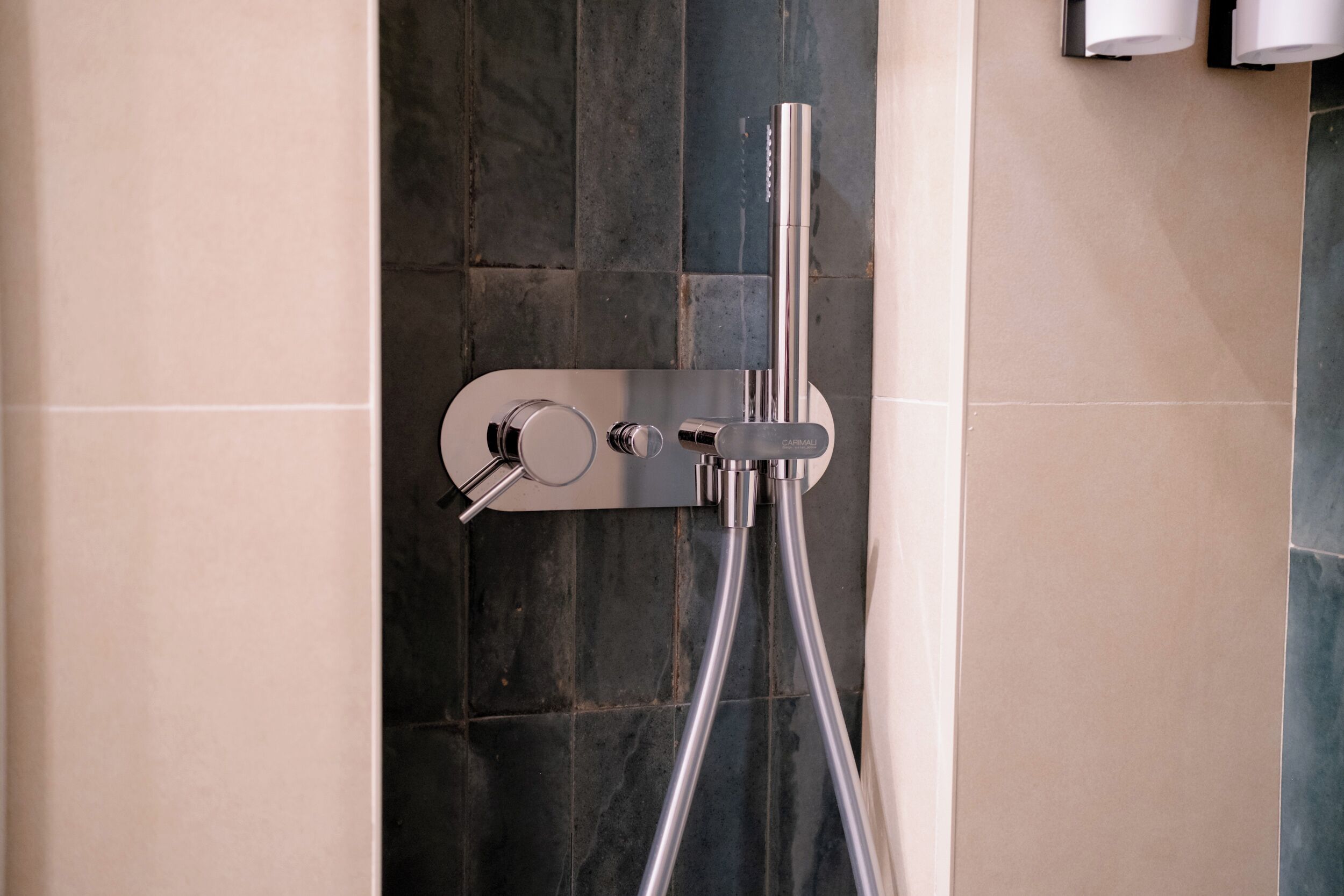 A modern shower with a handheld showerhead and a fixed showerhead, mounted on a wall with a mix of beige and dark tiles.