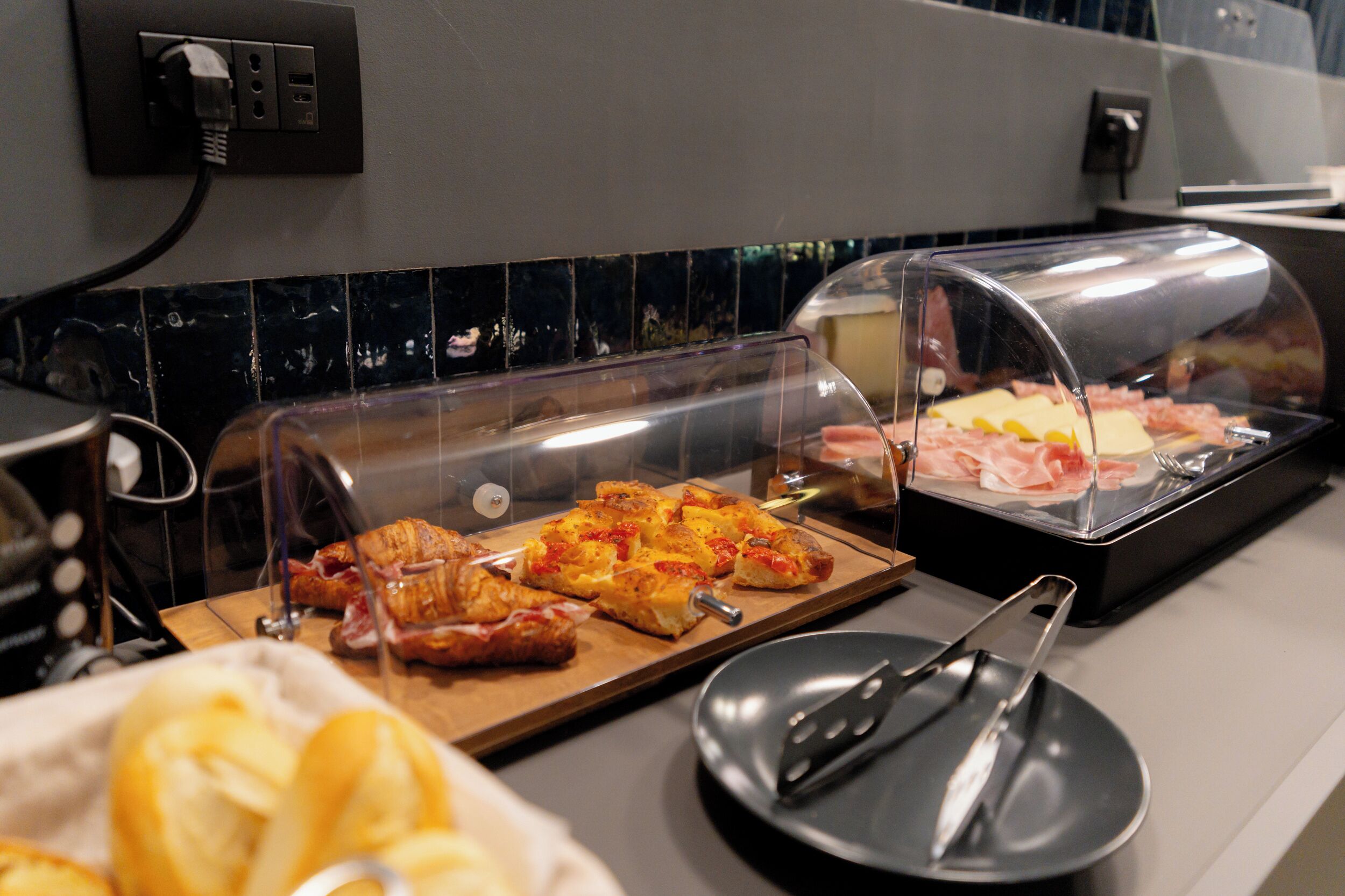 A buffet area with various food items, including a covered section with sliced meats and cheeses, and a tray with bread and a black plate with utensils.