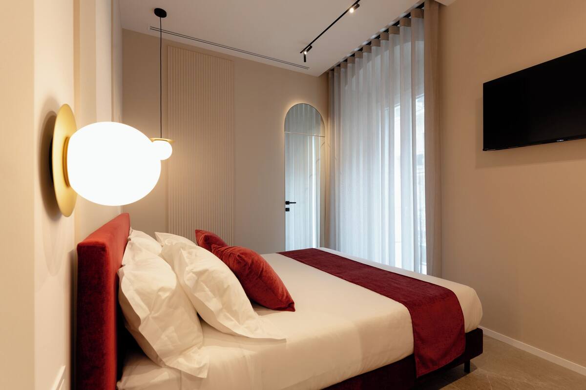 Una habitación de hotel moderna con una cama, un televisor montado y un amplio ventanal con cortinas.