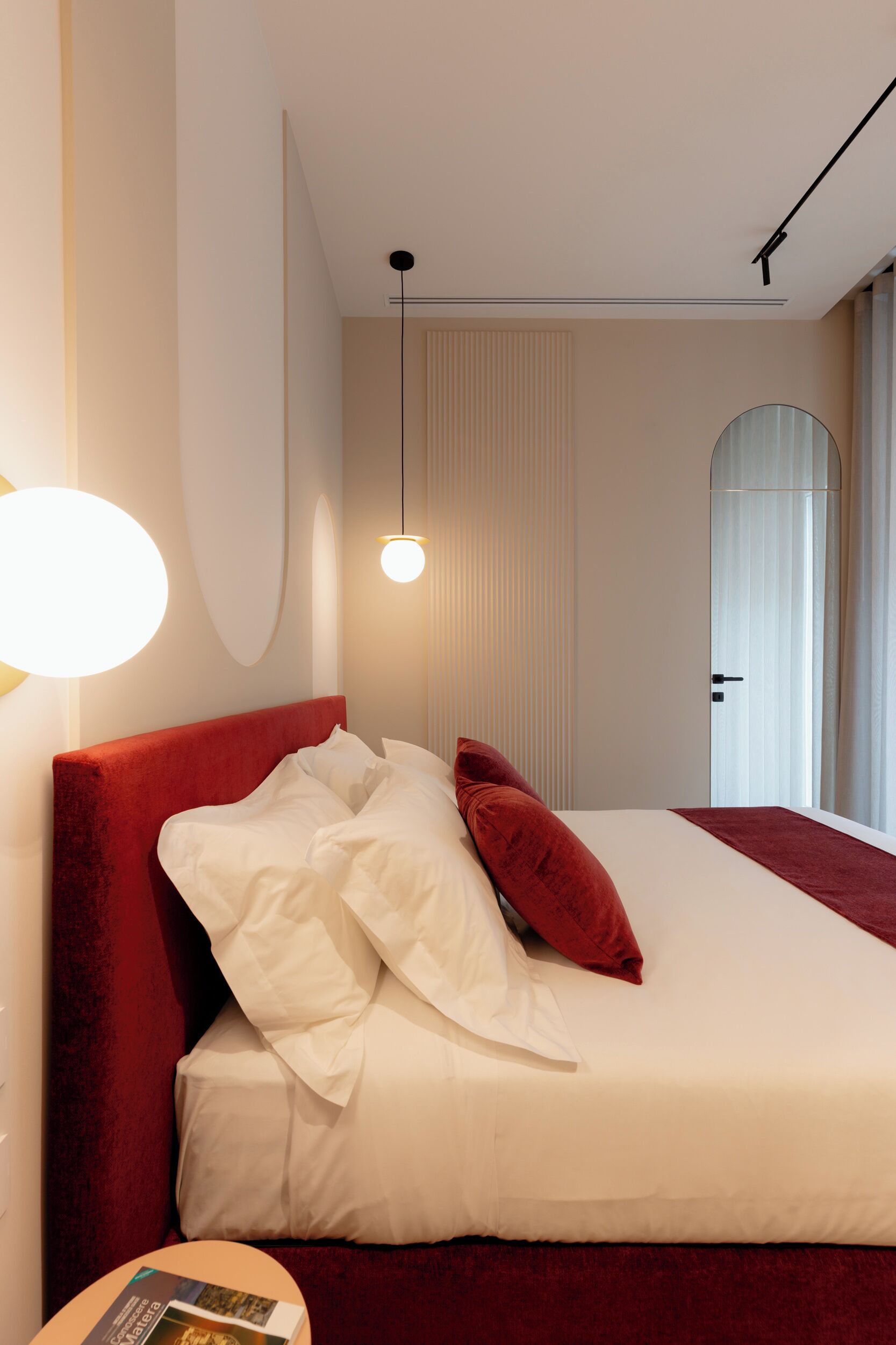 A hotel room with a large bed, a red headboard, white bedding, and a round wall light.