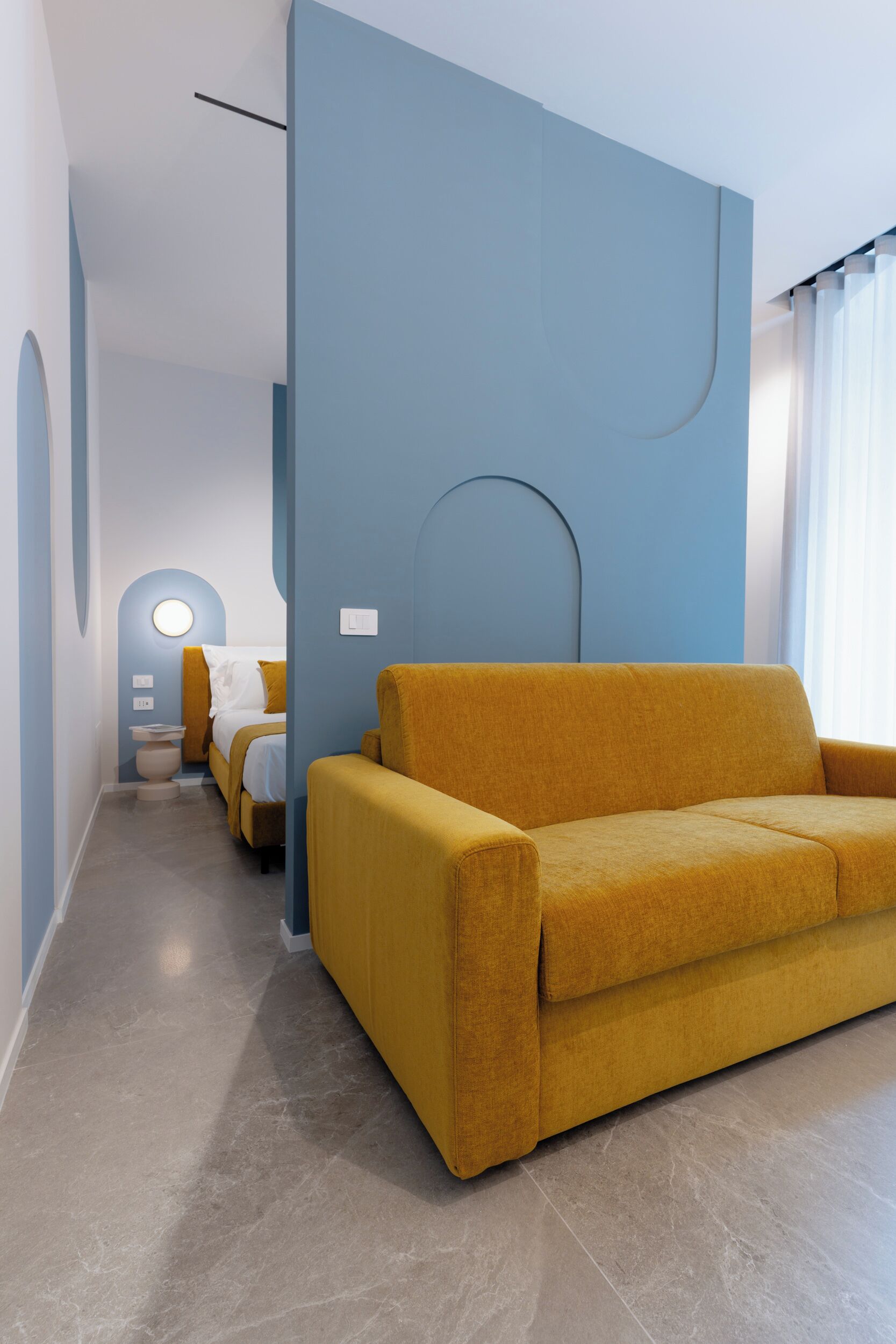 Una habitación de hotel moderna con un sofá amarillo, una cama con ropa blanca y una pared de acento azul.