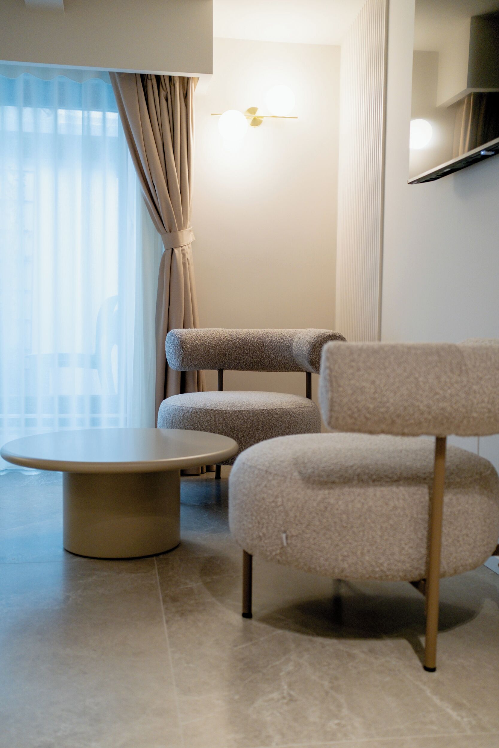 A modern room with a round table and two armchairs, a large window with curtains, and a wall-mounted light fixture.