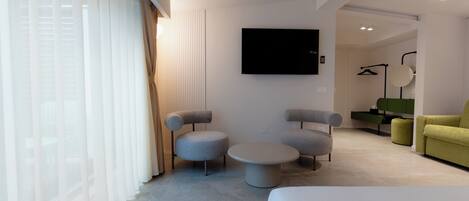 Ein modernes Hotelzimmer mit einem Bett, zwei Sesseln, einem runden Couchtisch und einem Fernseher an der Wand.
