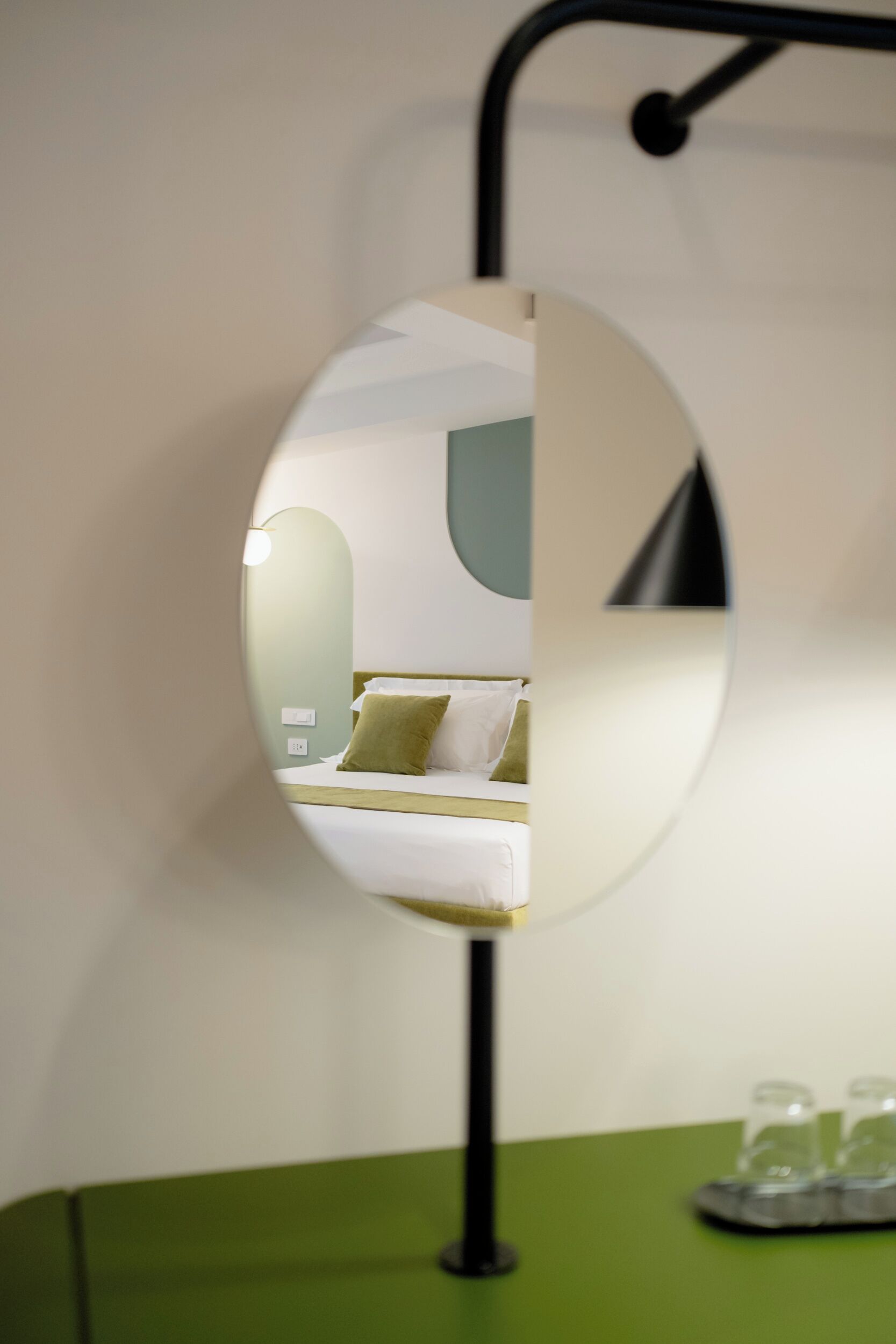 Un miroir reflétant un lit avec un oreiller vert, une table de chevet avec des lunettes, et un mur vert.