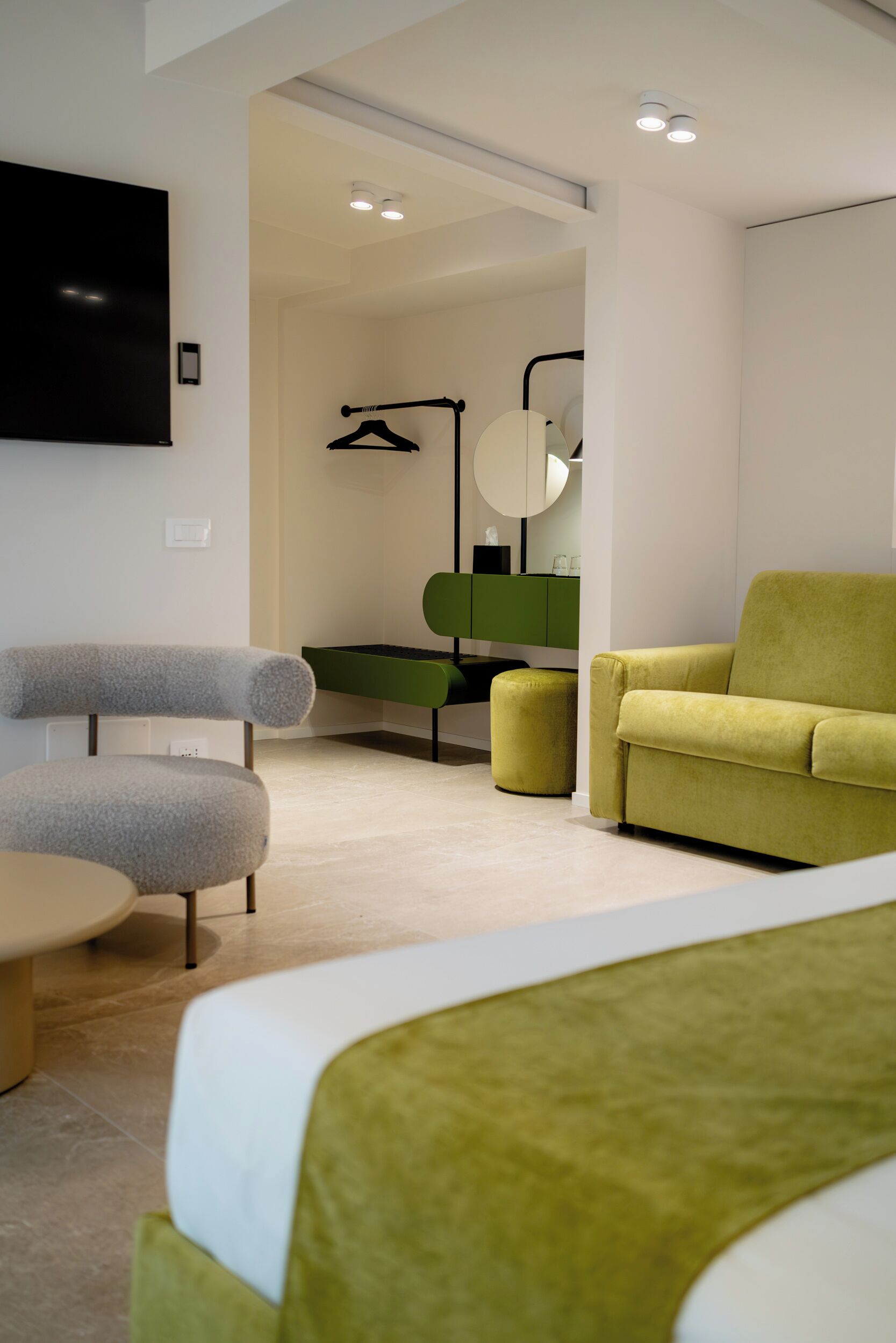 Habitación de hotel moderna con sofá verde, sillón gris, una pequeña mesa redonda y un reposapiés verde. Hay un televisor de pantalla plana montado en la pared y un mueble verde con un espejo encima.