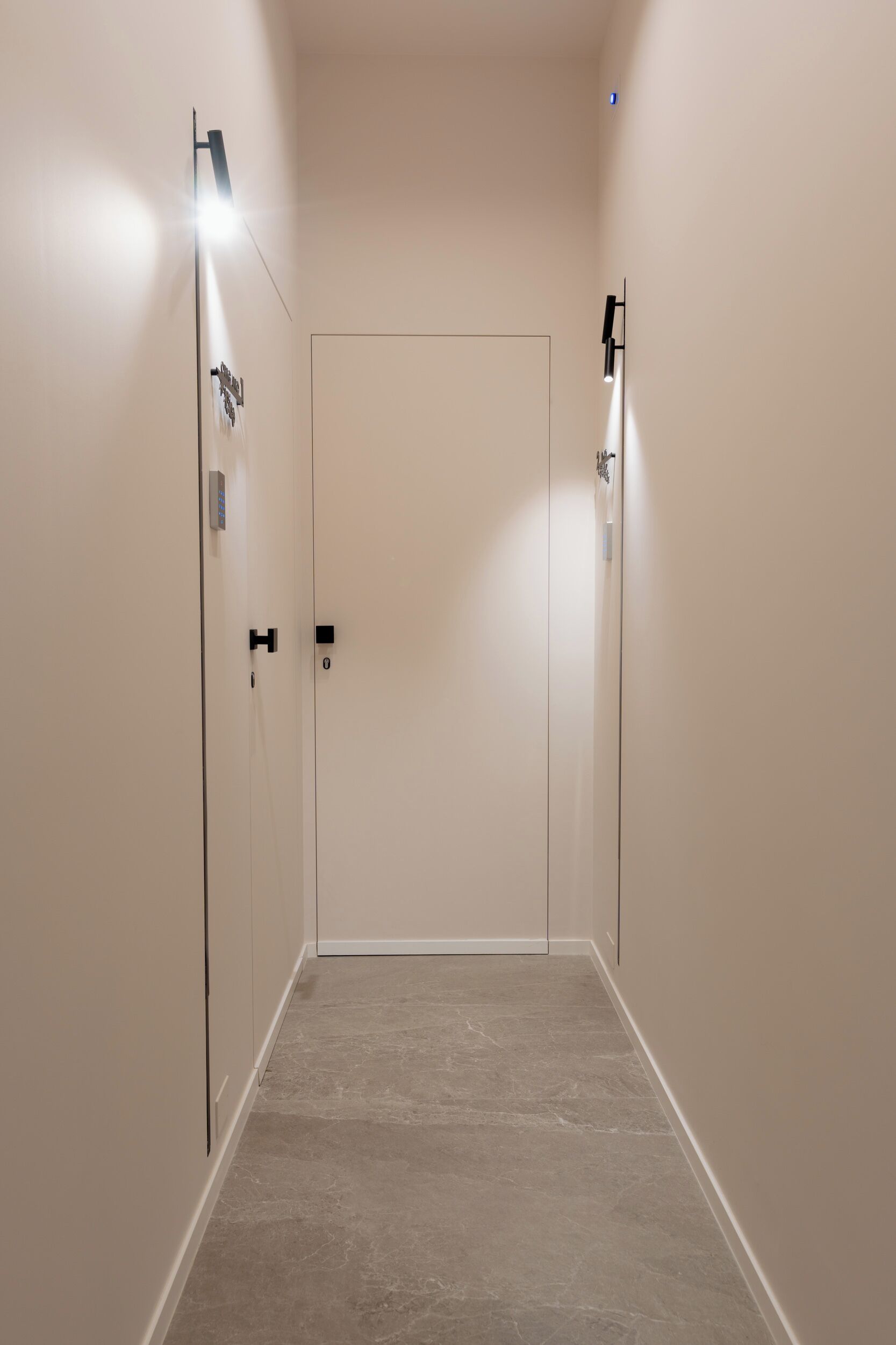 A narrow hallway with a door at the end, light fixtures on the walls, and a tiled floor.