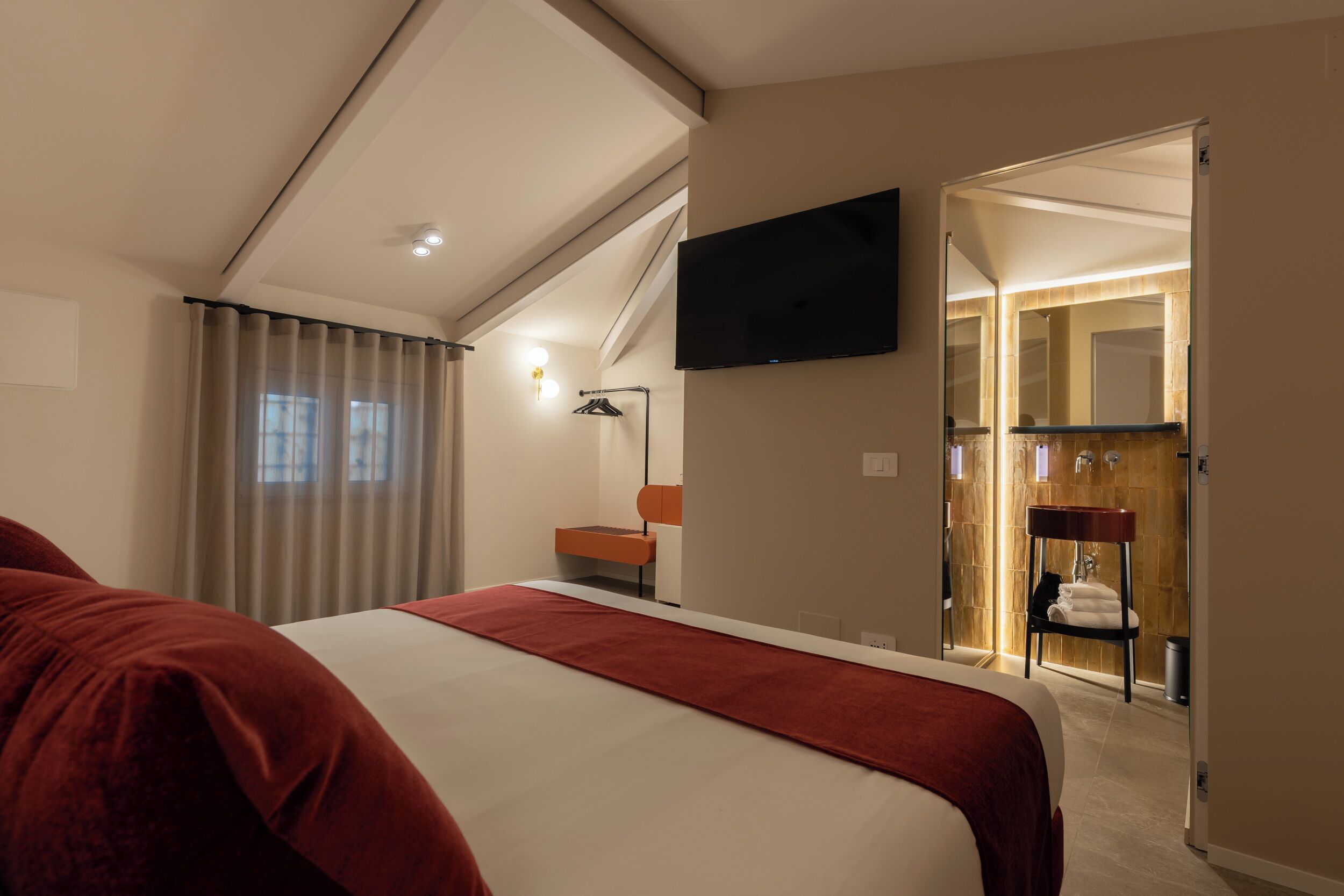 A hotel room with a large bed, a TV mounted on the wall, a bathroom with a mirror and sink, and a staircase leading to an upper level.