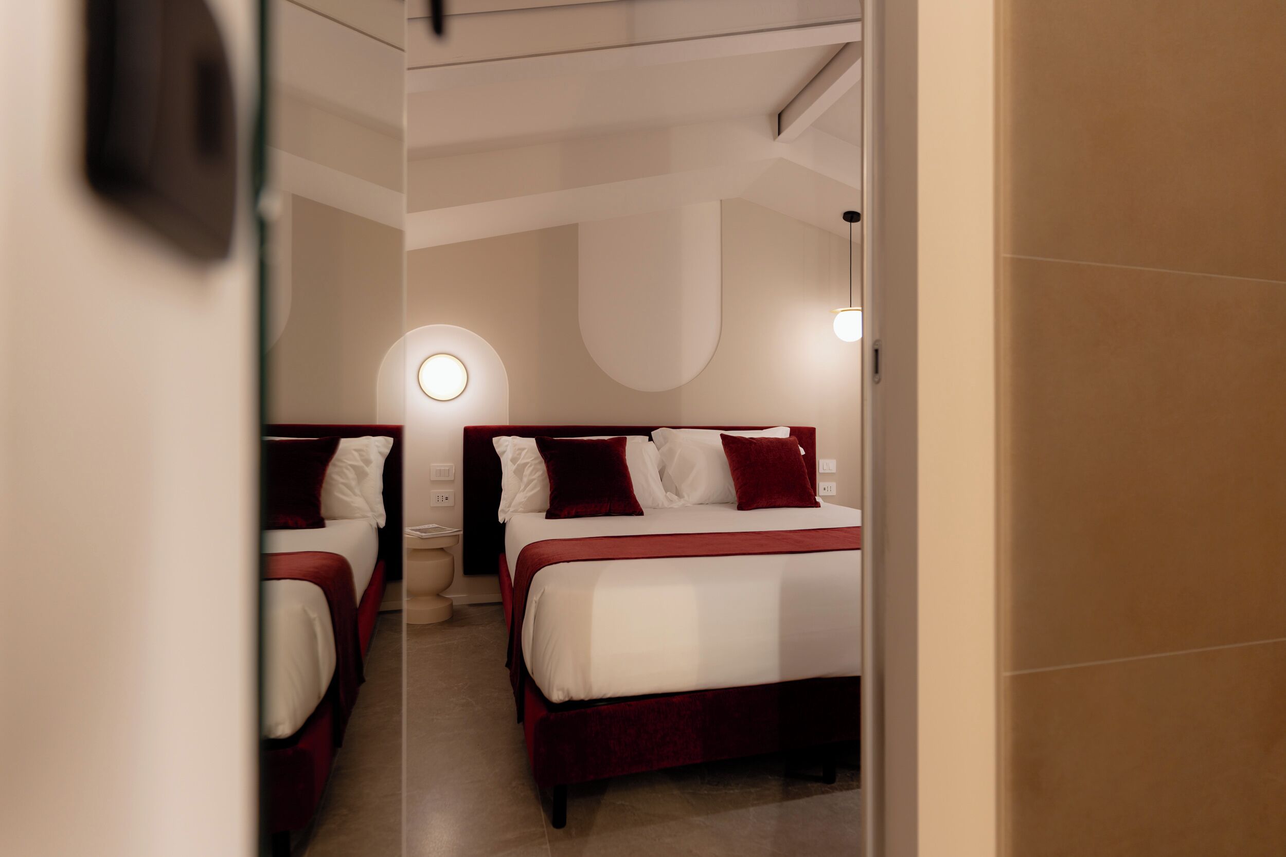 A hotel room with two beds, each with white linens and red accents, a round wall-mounted light, and a neutral-toned floor.