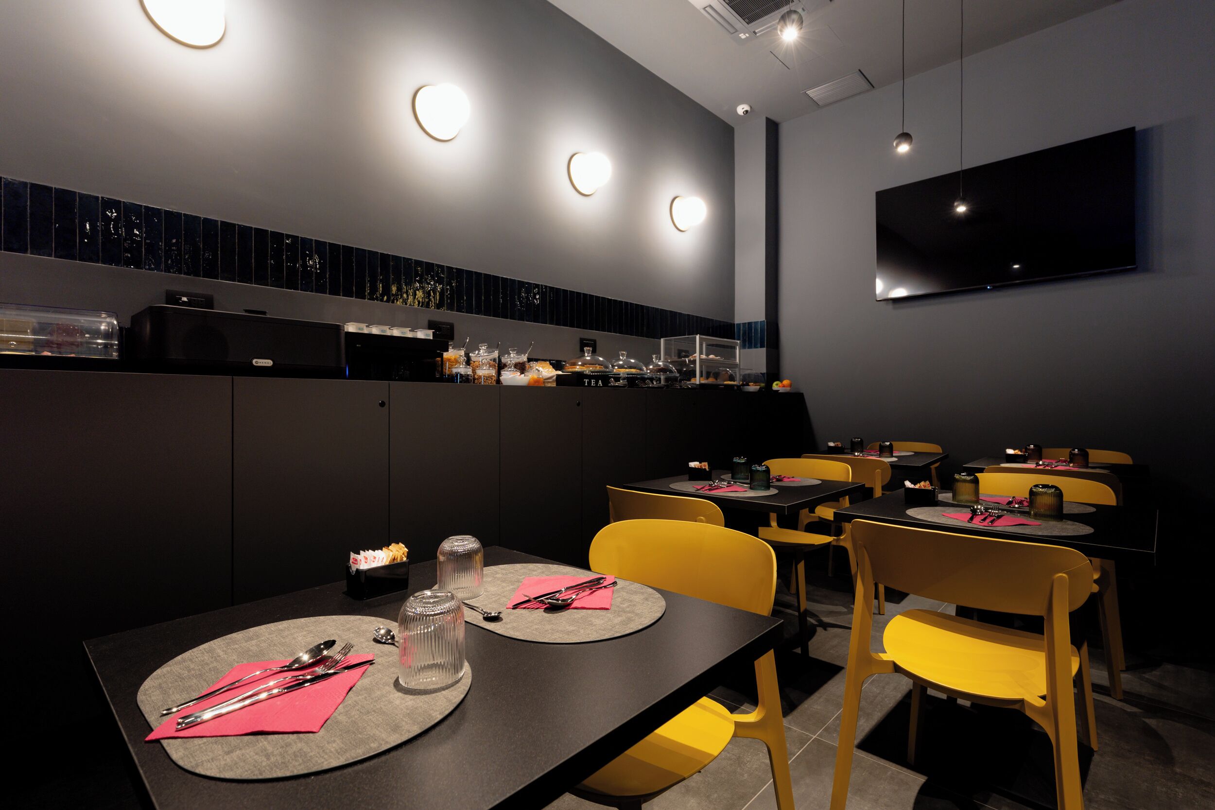 A modern dining area with tables set for two, equipped with cutlery and napkins, and a flat-screen TV mounted on the wall.