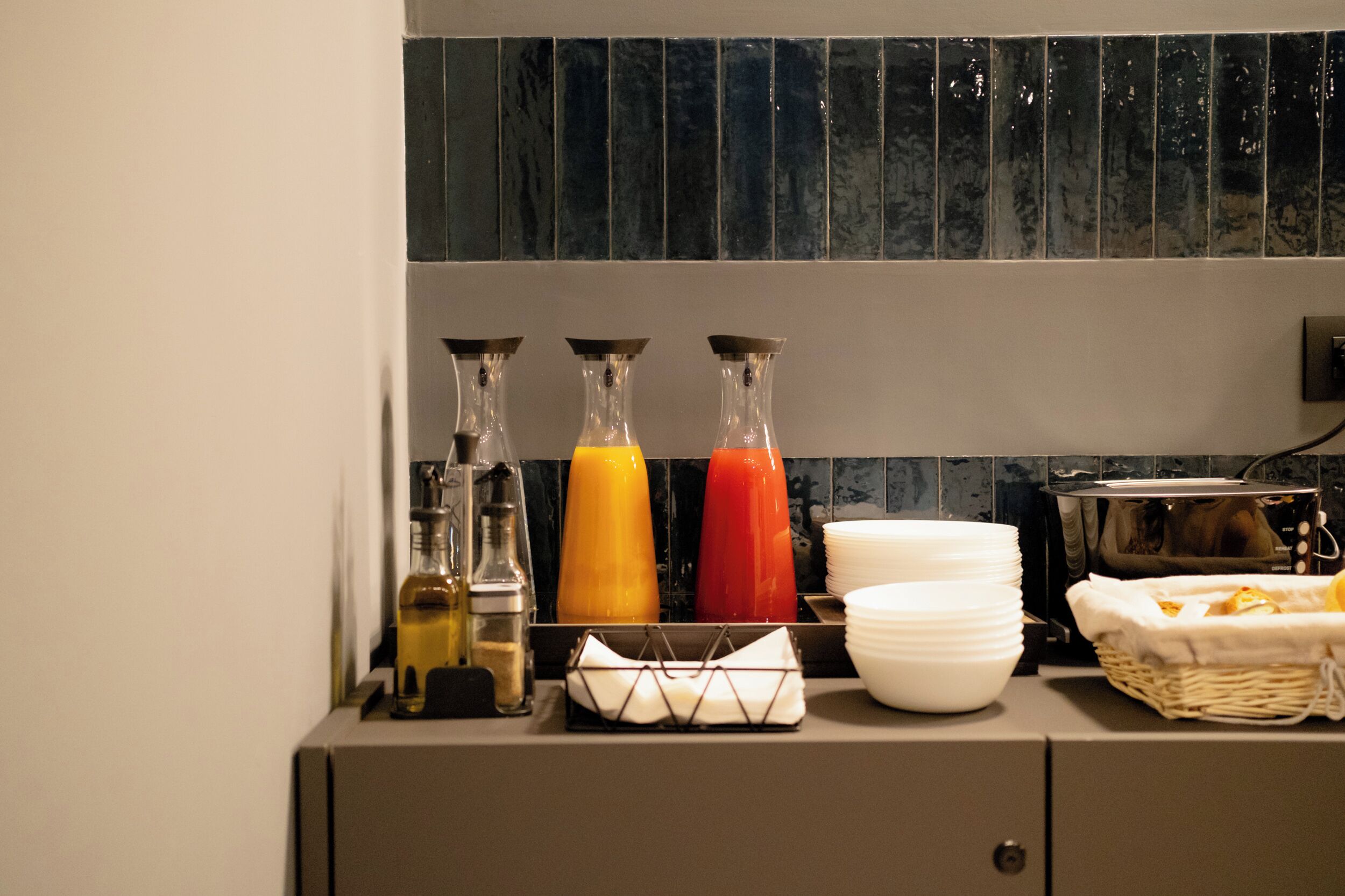 A breakfast bar with juice dispensers, stacked white plates, and a basket of bread.