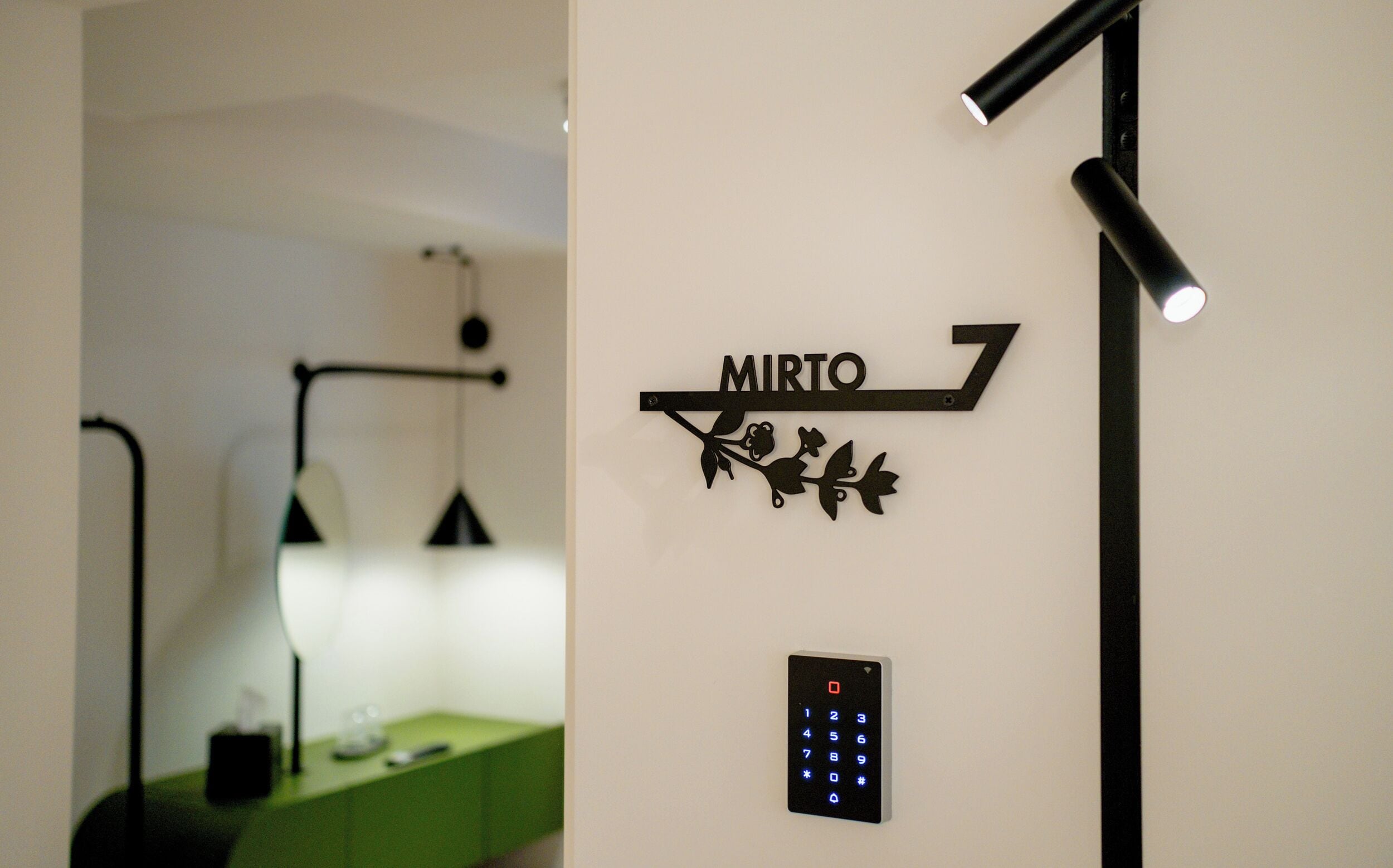 Eine an der Wand befestigte Schilder mit dem Schriftzug „MIRTO“ und einem dekorativen Element, das einen Zweig mit Blättern darstellt. Darunter befindet sich ein schwarzes Bedienfeld für den Aufzug.