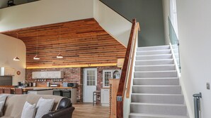 A spacious living area with a leather sofa, a kitchen with brick walls, and a staircase with a glass railing. - The Old Chapel (Berwick-upon-Tweed)