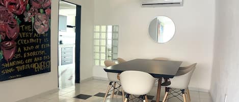 Un comedor con mesa y sillas, un ventilador de techo, un equipo de aire acondicionado, un espejo y una pared decorativa con una cita.