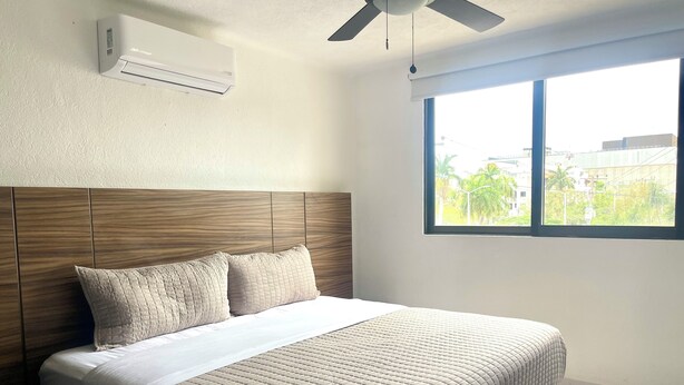 Une chambre à coucher comprenant un lit, un ventilateur de plafond, un climatiseur et une fenêtre donnant sur des bâtiments et de la verdure.
