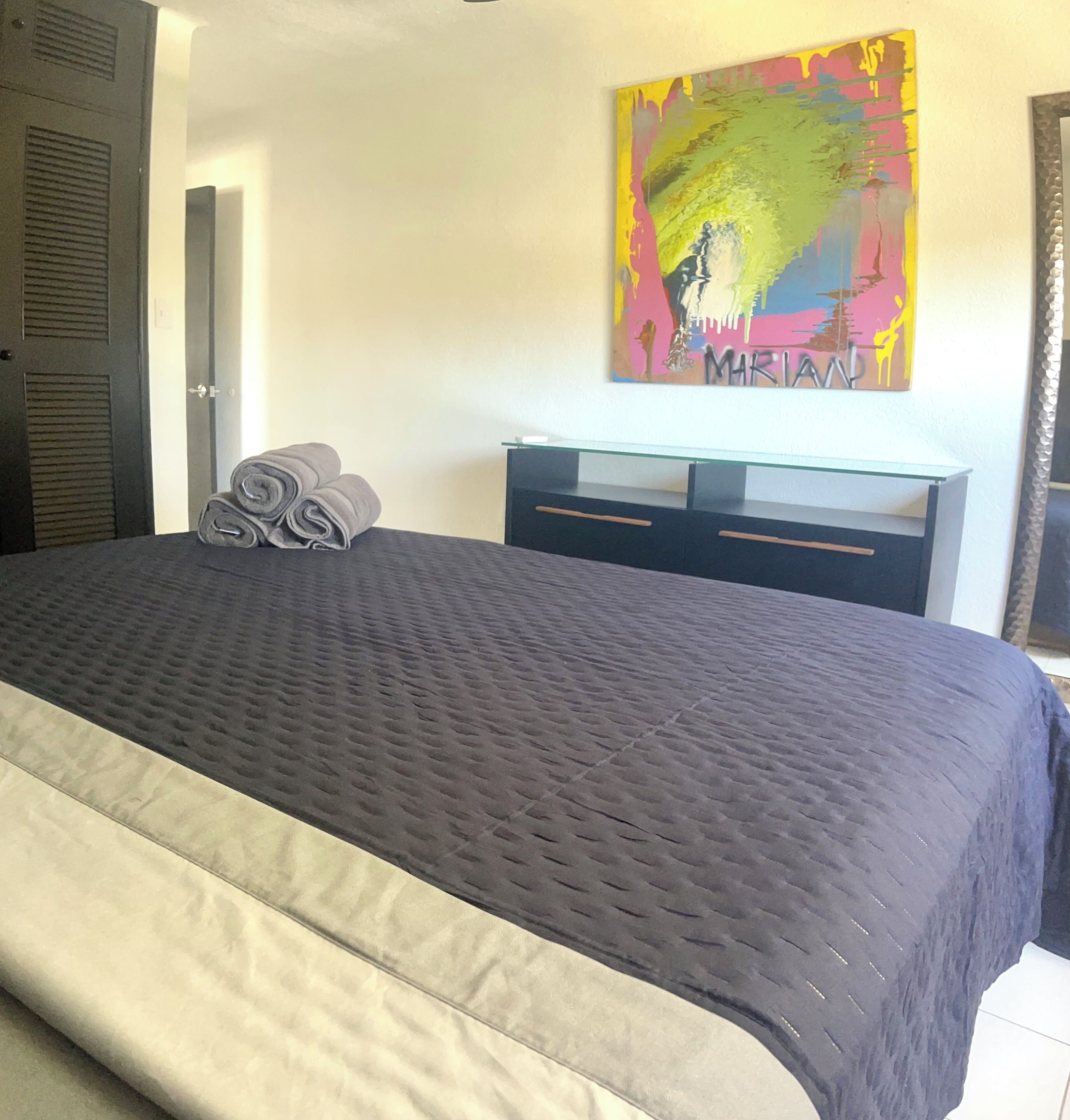 Un letto rifatto con cura, una coperta scura, un comodino in vetro, una moderna cassettiera e un quadro astratto dai colori vivaci appeso alla parete.