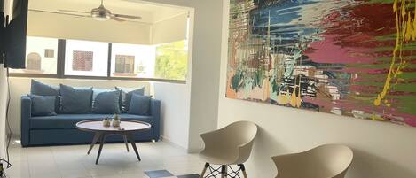 A modern living room with a blue sofa, two white chairs, a round coffee table, and a large abstract painting on the wall.