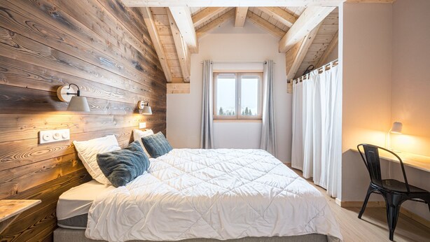 Un dormitorio con pared de madera, cama con ropa de cama blanca, ventana con cortinas y una silla.