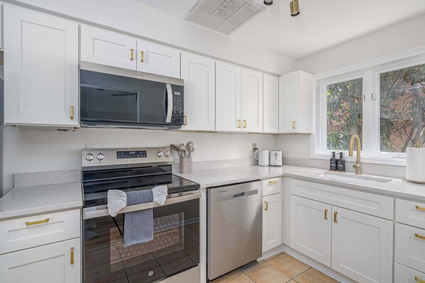 A modern kitchen with white cabinets, a built-in microwave, oven, and dishwasher, a window with a view of greenery, and a countertop with a kettle and soap dispensers.
