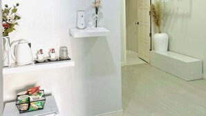 A modern, minimalist hotel room with a white color scheme, a built-in refrigerator, a small kitchen area, and a seating area with a bench and a vase of flowers.