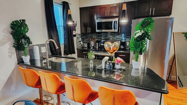Una cocina moderna con una isla central, armarios de madera oscura, electrodomésticos de acero inoxidable y taburetes de bar color naranja.