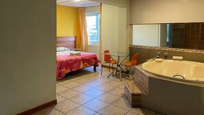 Ein Hotelzimmer mit Bett, einem Glastisch mit Stühlen, einer großen Badewanne und einem Fenster mit Vorhängen.