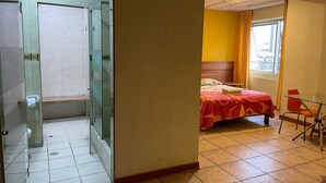 Ein Hotelzimmer mit Duschkabine aus Glas, einem Bett mit roter Bettdecke, einem Holzkopfteil, einem kleinen Tisch mit Stuhl und einem Fenster mit Vorhängen.
