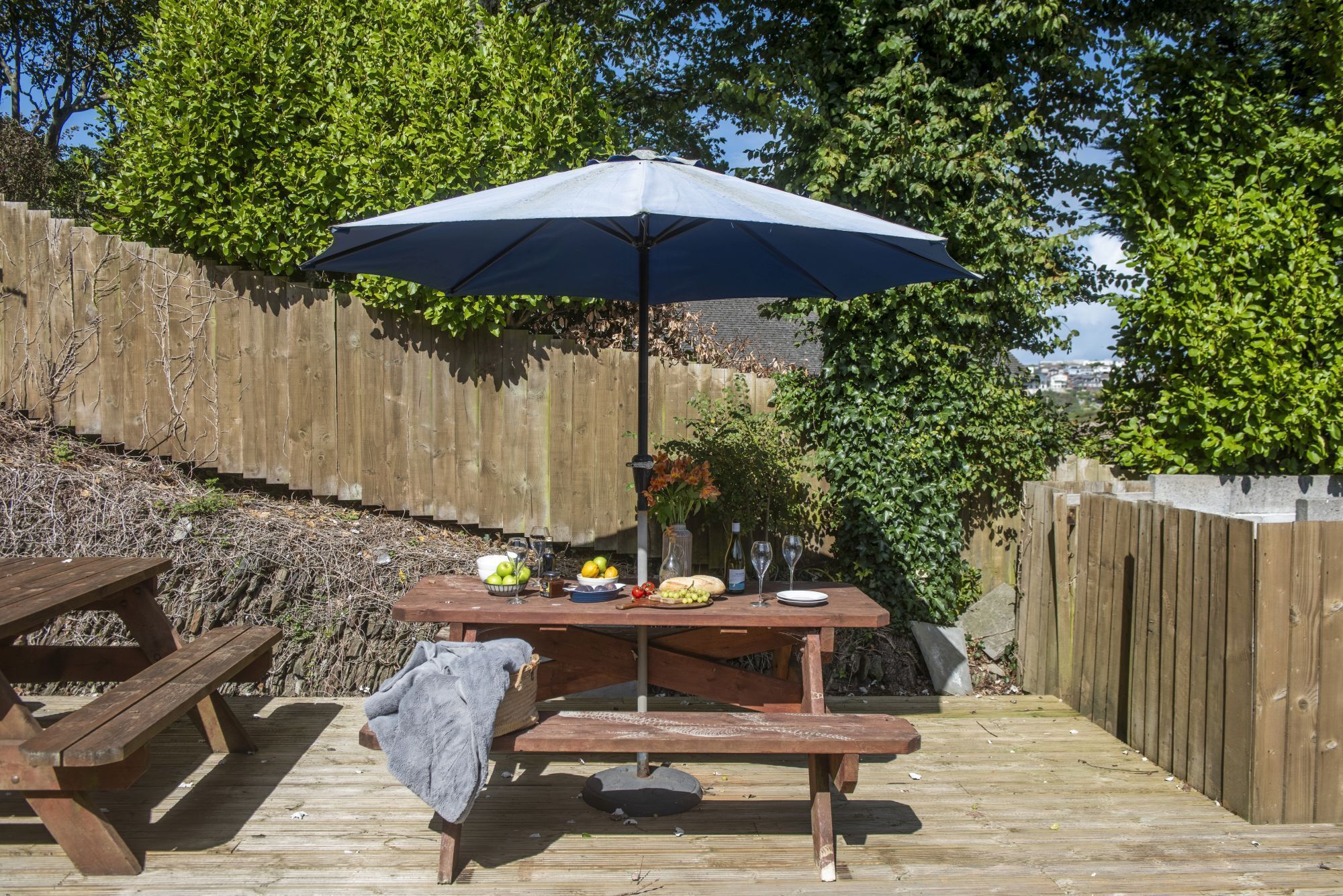 A wooden deck with a picnic table under a blue umbrella, surrounded by a wooden fence and greenery.