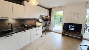 A modern kitchen with white cabinets, a black countertop, and a pendant light. There is a TV on a stand with a logo, and a door leading to a patio area.