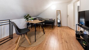 A small, modern room with a wooden floor, a desk with a chair, a sofa, a TV on a stand, and a mirror.
