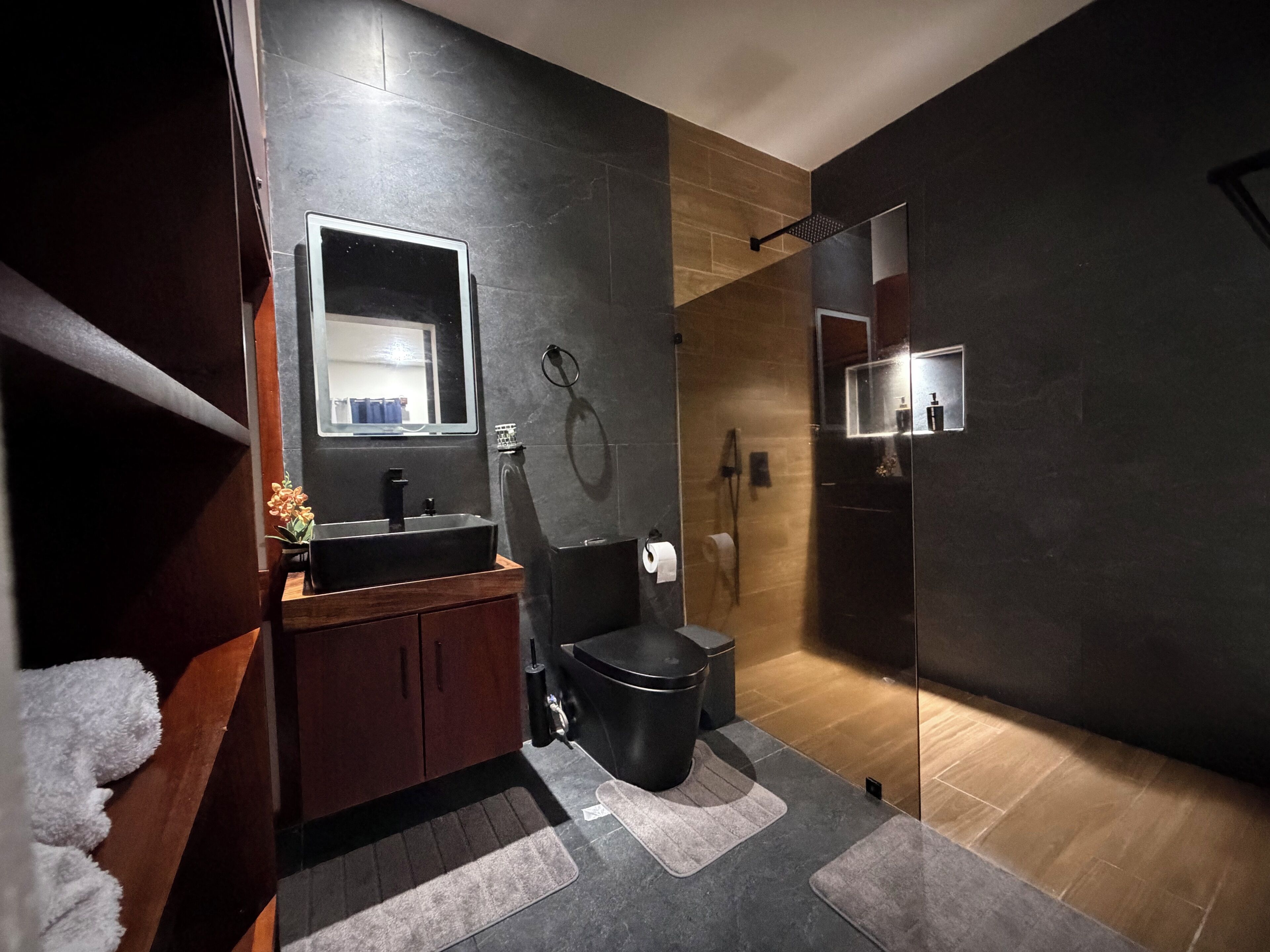 Une salle de bain moderne avec un mur recouvert de carreaux foncés, un meuble-lavabo en bois, des toilettes noires et une cabine de douche en verre.