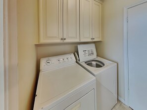 A compact laundry room with a front-loading washer and dryer, beige cabinets above, and a white door to the right.