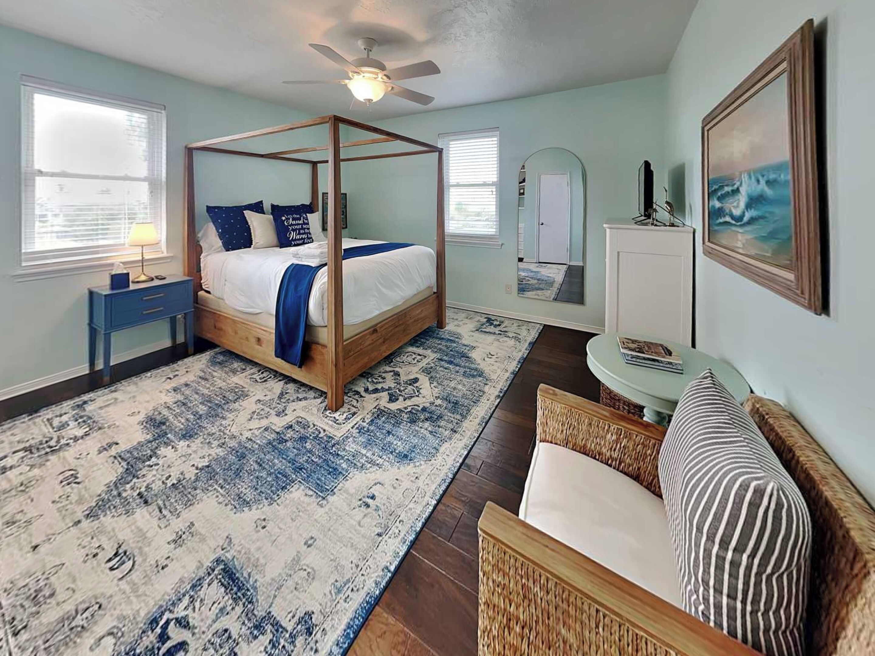 A four-poster bed with a ceiling fan, a blue and white patterned rug, a wicker chair, and a framed picture on the wall.
