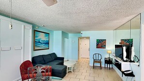 A living room with a sofa, a glass coffee table, and a dining table with chairs. There is a TV and a small table with a lamp. The room has light blue walls and a tiled floor. - The Salty Fountain by Distinctive Beach Rentals (Daytona Beach)