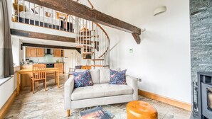Uma sala de estar espaçosa com sofá, mesa de centro e um banquinho. Uma escada de madeira leva a um andar superior com cozinha.