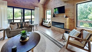 A spacious living room with a fireplace, wooden walls, and large windows offering a view of the outdoors.