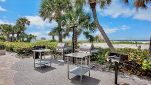 An outdoor patio with two barbecue grills, a table, and chairs, surrounded by palm trees and a view of the beach. - Peppertree #507- Private Access (Siesta Key)
