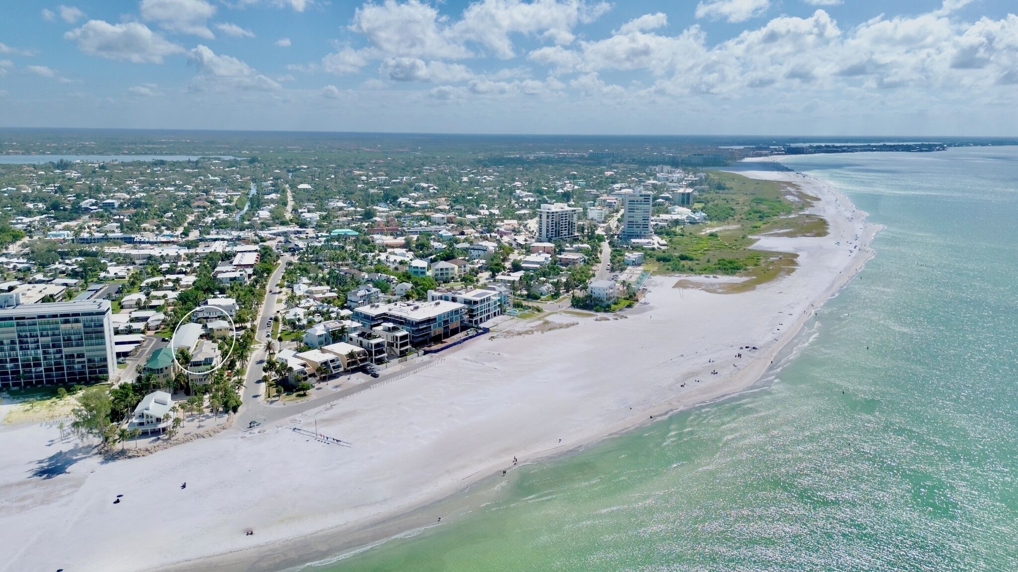 An aerial view of a coastal town with a wide sandy beach, a marina, and a mix of residential and commercial buildings.