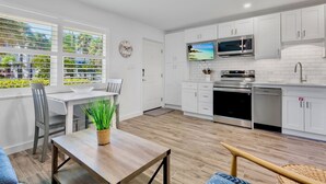 Een moderne keuken met witte kasten, een magnetron, oven en vaatwasser. Er is een eethoek met een tafel en stoelen, en een uitzicht op groen door het raam.