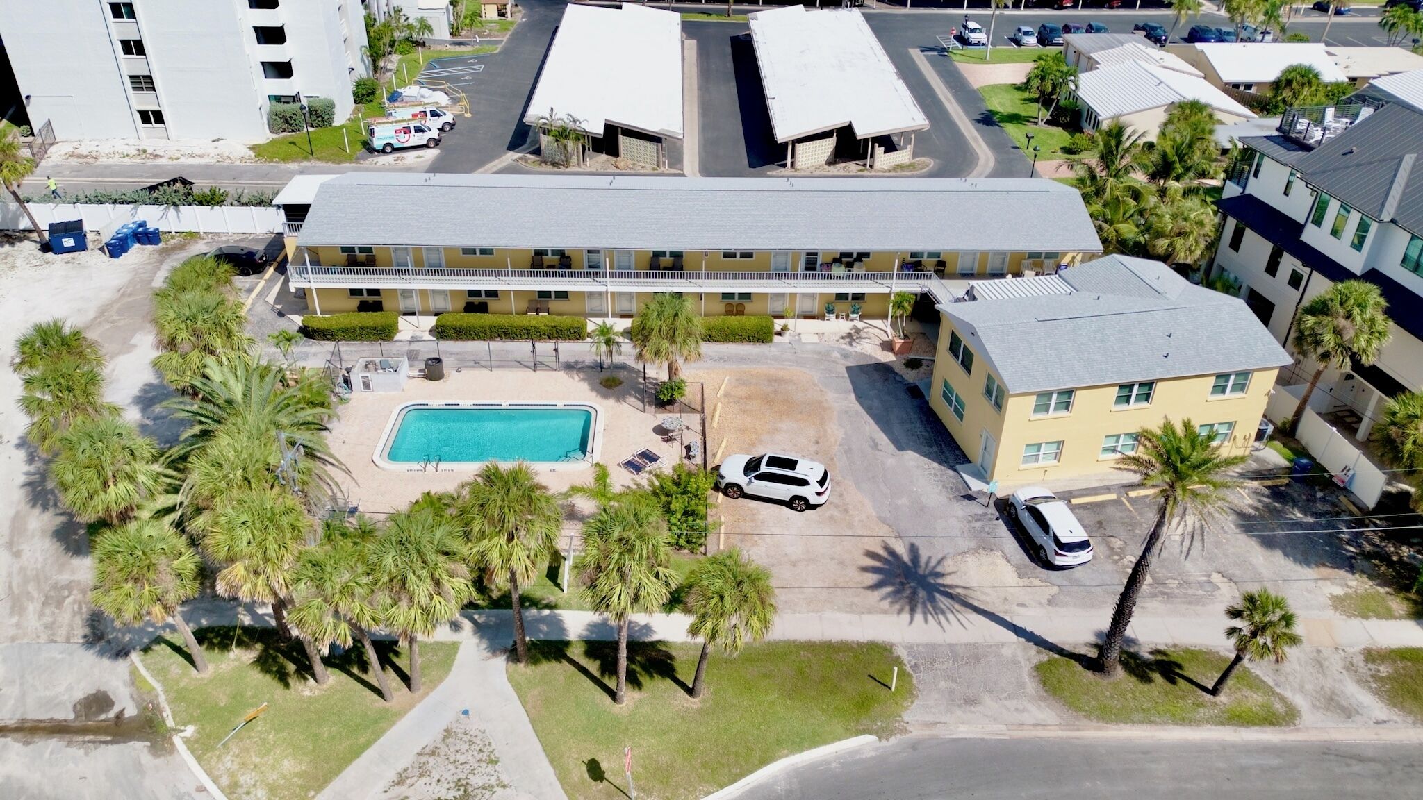 Vista aérea de un hotel con piscina, múltiples espacios de estacionamiento y palmeras circundantes.