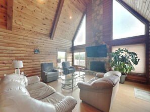 A log cabin interior with a stone fireplace, leather sofas, and a large window offering a view of the outdoors. - Tranquil Cove by Jackson Mountain Rentals (Sevierville)