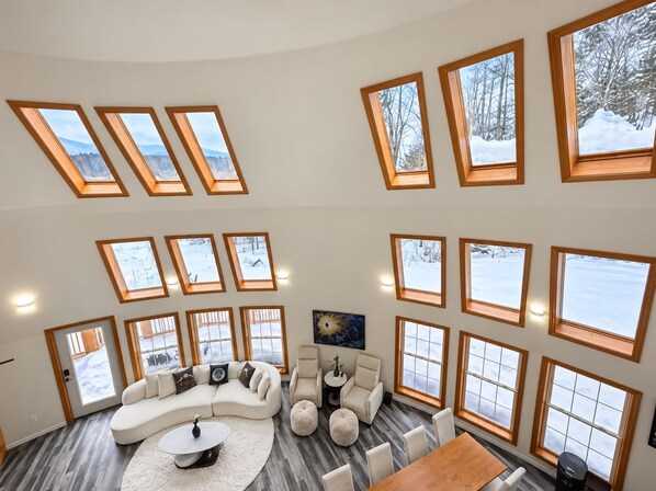 A spacious living room with a large sectional sofa, a round coffee table, and a dining table. The room has multiple skylights and large windows with views of a snowy landscape.