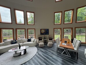 A spacious living room with a large sectional sofa, a coffee table, and a dining table with chairs. There are multiple windows providing views of the outdoors.