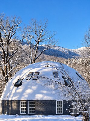 A snow-covered house with a dome roof, surrounded by bare trees and mountains in the background.